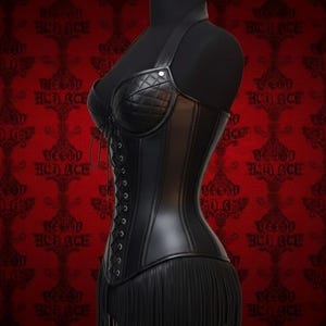 May include: Black leather corset with a halter neck and fringe detail. The corset features a quilted bust and a lace-up back. The garment is displayed on a black mannequin against a red patterned background.