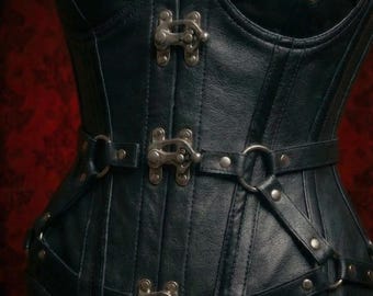 May include: Black leather corset with metal clasps and ring details. The corset features a structured design with multiple straps and rivets, creating a gothic or steampunk aesthetic. The garment is designed to cinch the waist.