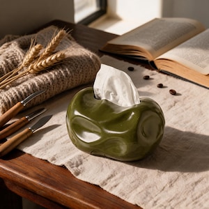 May include: An olive green ceramic tissue box cover with an irregular, rounded, and dimpled surface. The container features a central opening from which a single white paper tissue protrudes, providing a functional and sculptural accent.
