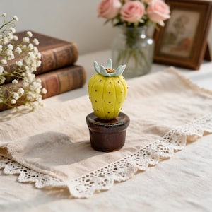 May include: A yellow ceramic cactus figurine with small brown dots and a light blue flower on top, set in a dark brown cylindrical base. The cactus has a rounded, ribbed shape and sits upright as a decorative object on a flat surface.
