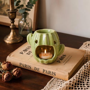 May include: A light green ceramic wax warmer shaped like a cactus with a central opening containing a small lit tea light candle. The cactus features two side arms and small circular cutouts, resting on a book titled PARIS DE PARIS.