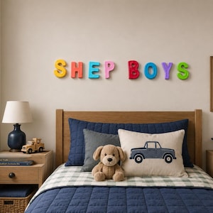 May include: A bedroom featuring a bed with a navy blue quilt, a beige pillow with a blue truck graphic, and a brown plush dog. Above the bed, colorful wall letters spell SHEP BOYS. A framed print reads LITTLE MAN with a green truck graphic.
