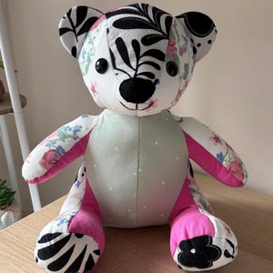 May include: A stuffed teddy bear constructed from various fabric patches. The head features a white background with black leaf patterns and small pink and blue flowers. The torso is light green with white dots, while the limbs feature pink accents.