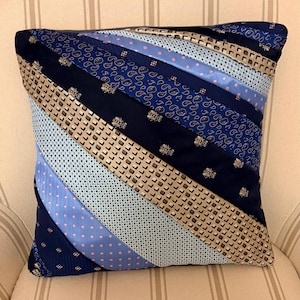 May include: A square throw cushion constructed from diagonal strips of various neckties in shades of navy blue, light blue, and tan. The fabric features patterns including small bee motifs, paisley swirls, polka dots, and a grid-like geometric design.