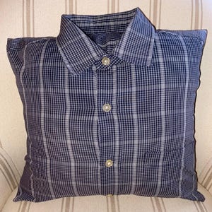 May include: A square throw cushion constructed from a navy blue and white plaid button-down shirt. The cushion features a pointed collar at the top, a central vertical placket with three visible light-coloured buttons, and a single chest pocket on the right.