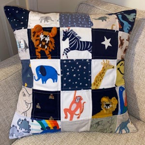 May include: A square patchwork cushion featuring appliquéd animals such as a tiger, zebra, elephant, giraffe, monkey, and lion. The design incorporates various fabrics in blue, white, and neutral tones. The cushion is placed on a neutral-toned sofa.