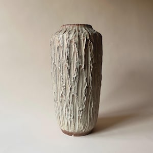 Può includere: Un vaso in ceramica alto e strutturato, di forma cilindrica. Il vaso presenta un motivo verticale e scanalato in tonalità di bianco e marrone, creando una superficie tattile. La base e il bordo hanno una tonalità marrone più scura, aggiungendo contrasto.
