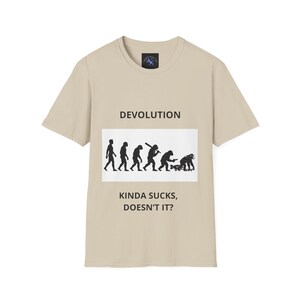 Può includere: T-shirt beige con la scritta "DEVOLUTION" sopra una grafica che raffigura l'evoluzione dell'uomo, che termina con una figura curva. Sotto la grafica, il testo recita "KINDA SUCKS, DOESN'T IT?"