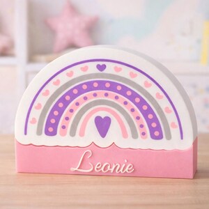 Könnte beinhalten: Ein rosa-weißes Regenbogen-Nachtlicht mit dem Namen "Leonie" auf der Vorderseite. Das Regenbogen-Design zeigt lila, graue und rosa Bögen mit Herz- und Punktdetails. Der Hintergrund umfasst einen rosa Stern und ein Stofftier.
