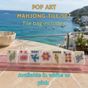 Pu&ograve; includere: Un set di sette tessere rettangolari bianche da mahjong disposte in fila su un supporto rosa. Ogni tessera presenta grafiche pop art colorate, tra cui fulmini, un aereo, un fiore, un gelato, stelle e una saponetta con la scritta SOAP.