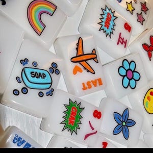 Pu&ograve; includere: Una collezione di tessere rettangolari bianche da mahjong con disegni grafici colorati che includono un arcobaleno, una saponetta blu, un aereo arancione, un fiore blu, un palloncino a forma di animale rosso, una tavolozza di colori e le scritte EAST e CRAK.