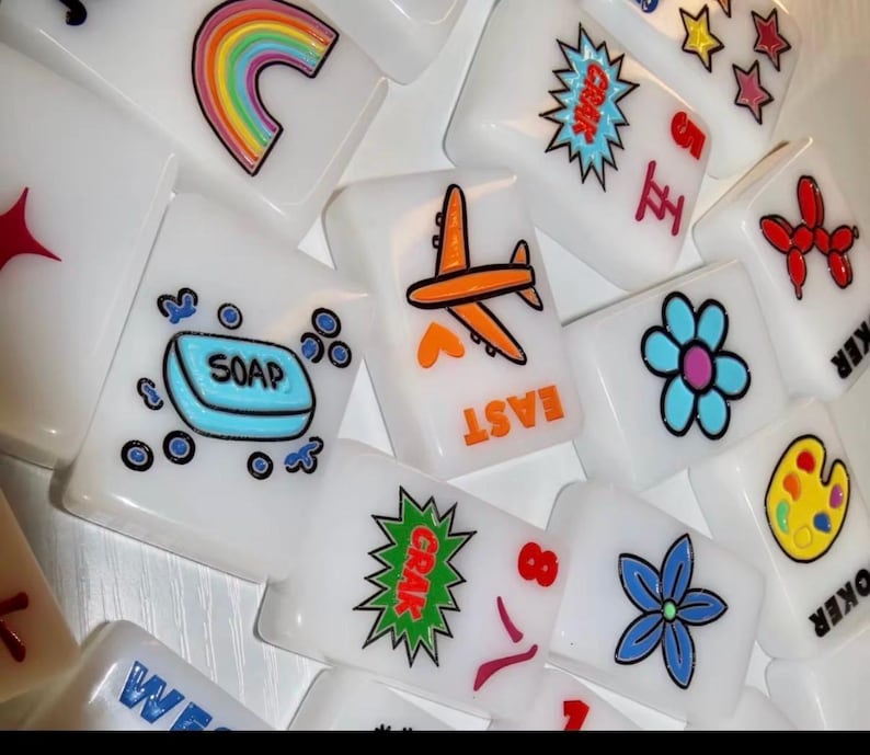 Pu&ograve; includere: Una collezione di tessere rettangolari bianche da mahjong con disegni grafici colorati che includono un arcobaleno, una saponetta blu, un aereo arancione, un fiore blu, un palloncino a forma di animale rosso, una tavolozza di colori e le scritte EAST e CRAK.