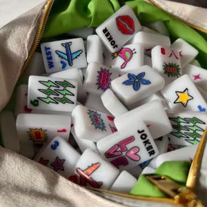 Pu&ograve; includere: Una collezione di tessere rettangolari bianche da Mahjong con disegni grafici colorati tra cui labbra, aerei, stelle, fiori, fulmini e il testo JOKER e LOVE, contenute in una borsa di tessuto beige con fodera interna verde brillante.