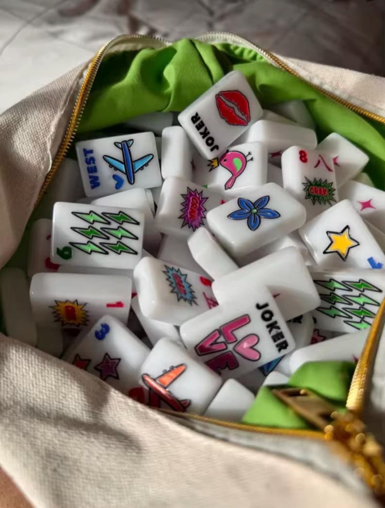 Pu&ograve; includere: Una collezione di tessere rettangolari bianche da Mahjong con disegni grafici colorati tra cui labbra, aerei, stelle, fiori, fulmini e il testo JOKER e LOVE, contenute in una borsa di tessuto beige con fodera interna verde brillante.