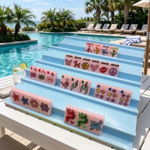 Pu&ograve; includere: Un supporto per mahjong a gradini azzurro contenente una collezione di tessere rosa con vari grafici colorati, tra cui aerei etichettati North, East, West, South, carte Joker, numeri, fiori, stelle, fulmini e un drago.