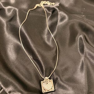 May include: Silver necklace with a square pendant. The pendant has a heart-shaped design filled with small, sparkling stones. The necklace is on a black satin surface.