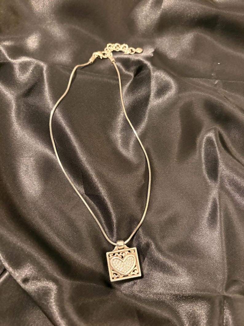 May include: Silver necklace with a square pendant. The pendant has a heart-shaped design filled with small, sparkling stones. The necklace is on a black satin surface.