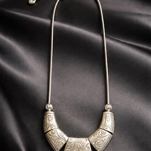 May include: Silver-toned necklace with an ornate, engraved pendant. The pendant is composed of three connected sections, each with detailed patterns. The necklace has a delicate chain and is displayed against a black satin background.