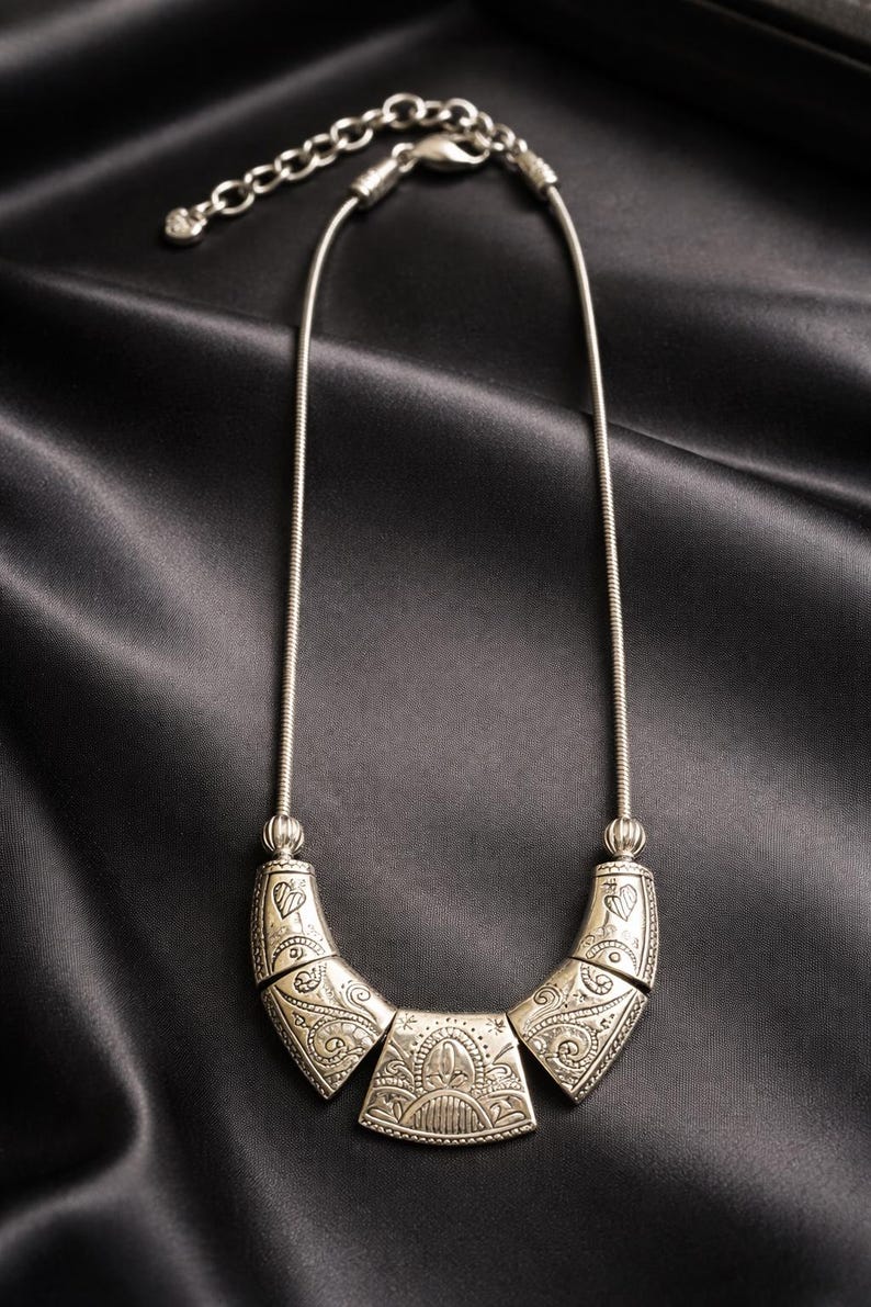 May include: Silver-toned necklace with an ornate, engraved pendant. The pendant is composed of three connected sections, each with detailed patterns. The necklace has a delicate chain and is displayed against a black satin background.