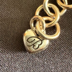 May include: Close-up of a gold-toned chain bracelet with large, circular links. A heart-shaped charm with an ornate design hangs from the chain. The background is a textured, dark brown surface.