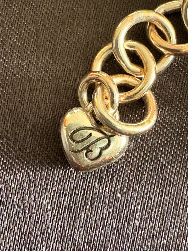 May include: Close-up of a gold-toned chain bracelet with large, circular links. A heart-shaped charm with an ornate design hangs from the chain. The background is a textured, dark brown surface.