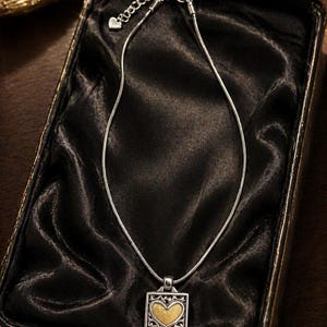 May include: Silver necklace with a square pendant featuring a gold heart design. The necklace is presented in a black jewellery box with a satin lining. The pendant has an ornate silver border.