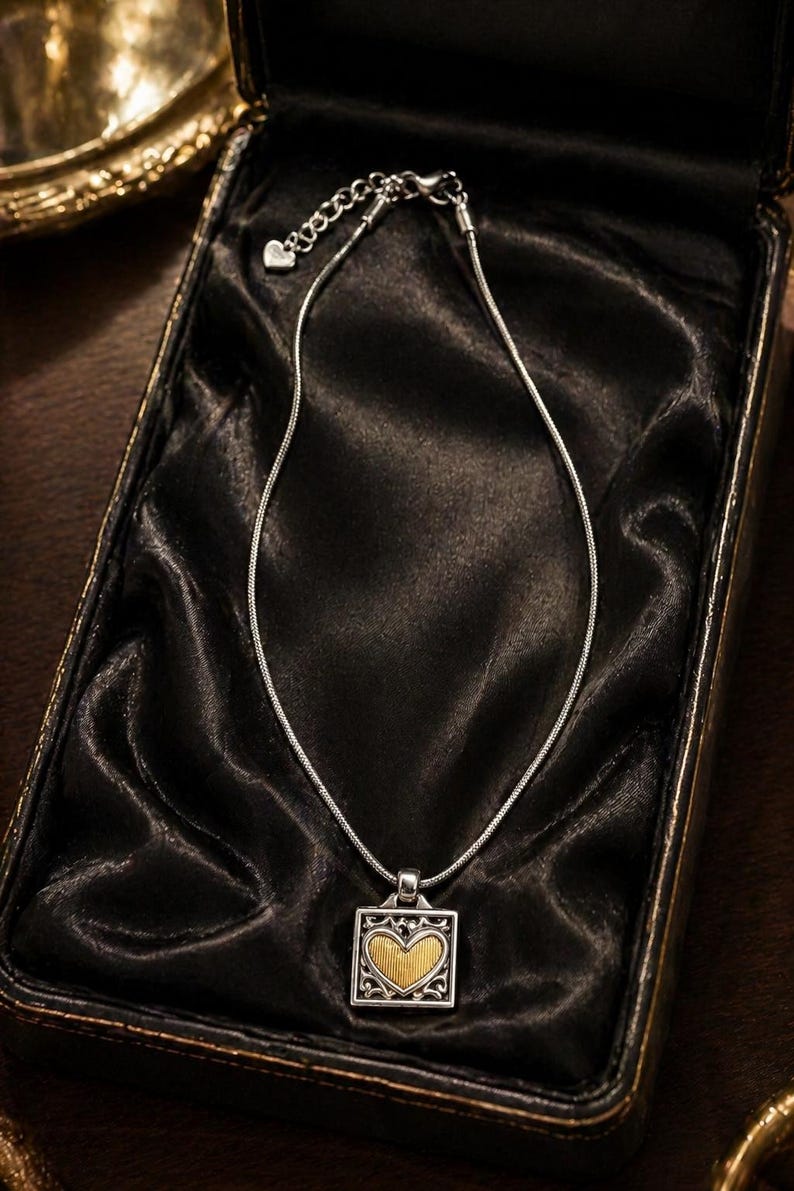 May include: Silver necklace with a square pendant featuring a gold heart design. The necklace is presented in a black jewellery box with a satin lining. The pendant has an ornate silver border.