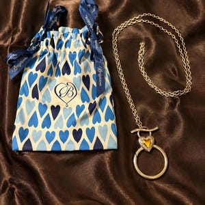 May include: A silver-toned necklace with a heart-shaped pendant featuring a yellow center stone and a circular design. It is accompanied by a drawstring pouch with a blue heart pattern and a ribbon with the word "Brighton".