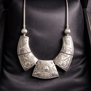 May include: Silver-toned necklace with an ornate, engraved design. The necklace features a central pendant and two curved side pieces, all with intricate patterns and a heart motif. The necklace is displayed in a black jewelry box.