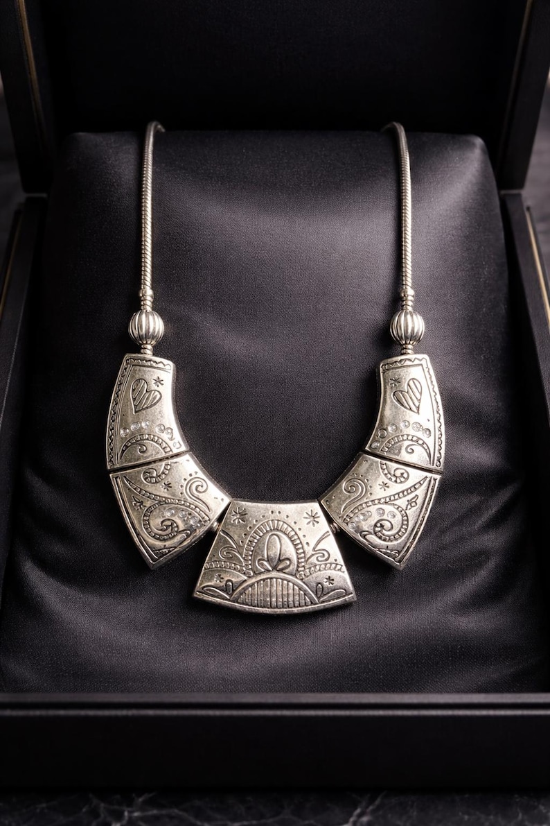 May include: Silver-toned necklace with an ornate, engraved design. The necklace features a central pendant and two curved side pieces, all with intricate patterns and a heart motif. The necklace is displayed in a black jewelry box.