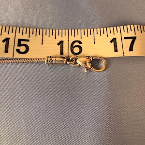 May include: A gold-colored necklace clasp with a heart-shaped design, measuring approximately 16 inches (40.6 cm) in length, is shown against a light blue background. The clasp is connected to a thin, silver chain, and a measuring tape is placed above it.