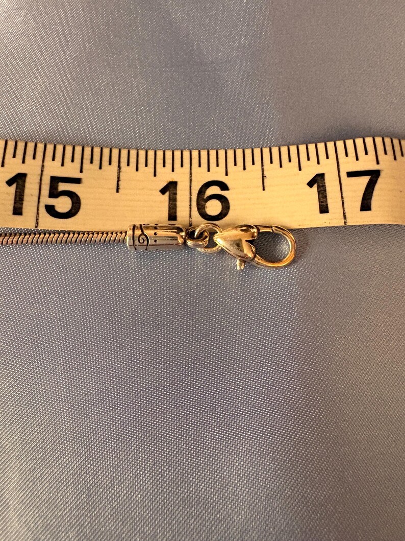 May include: A gold-colored necklace clasp with a heart-shaped design, measuring approximately 16 inches (40.6 cm) in length, is shown against a light blue background. The clasp is connected to a thin, silver chain, and a measuring tape is placed above it.