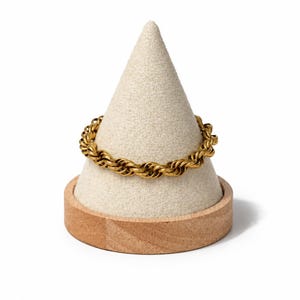 May include: A gold-toned rope chain bracelet featuring a twisted, interlocking link design. The bracelet is displayed on a beige, cone-shaped stand with a circular wooden base, highlighting the metallic texture and the continuous circular form.