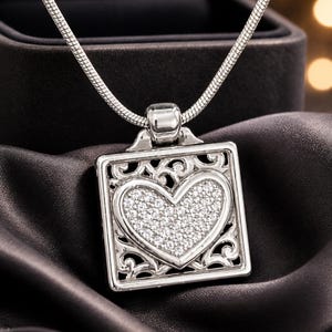 May include: A silver necklace with a square pendant. The pendant has a heart-shaped centre filled with small, sparkling stones, and an ornate, openwork border. The necklace is displayed on a dark, silky fabric.