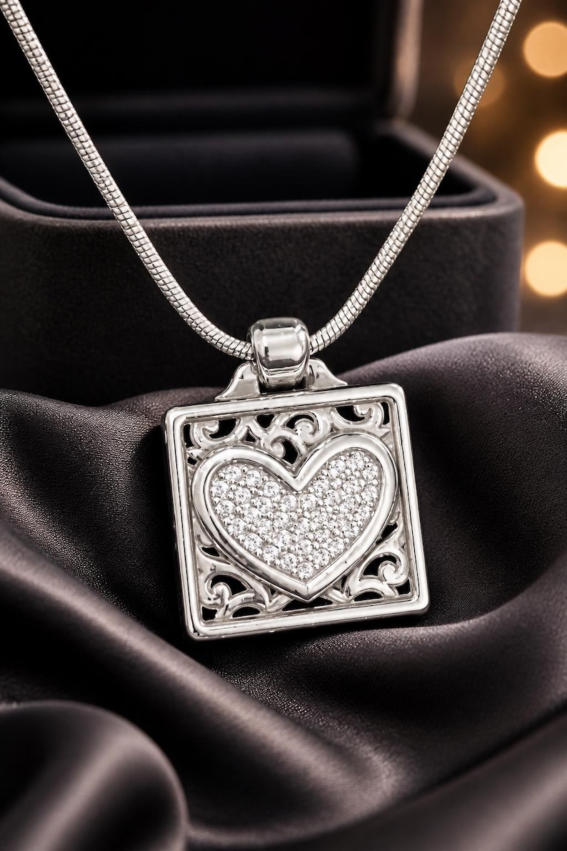 May include: A silver necklace with a square pendant. The pendant has a heart-shaped centre filled with small, sparkling stones, and an ornate, openwork border. The necklace is displayed on a dark, silky fabric.