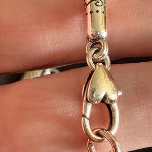 May include: Close-up of a silver necklace clasp. The clasp is heart-shaped and connects to a silver chain. The clasp is connected to a decorative silver tube with etched designs. The necklace is likely a fashion accessory.