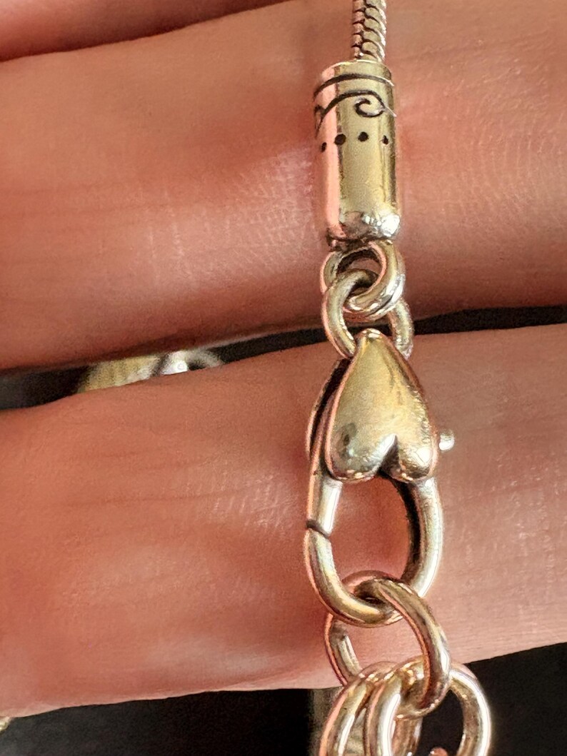May include: Close-up of a silver necklace clasp. The clasp is heart-shaped and connects to a silver chain. The clasp is connected to a decorative silver tube with etched designs. The necklace is likely a fashion accessory.