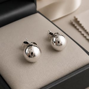 May include: A pair of polished silver ball stud earrings with a smooth, reflective spherical surface. Each earring features a standard post and butterfly back closure, showcasing a clean, metallic finish that catches the light.