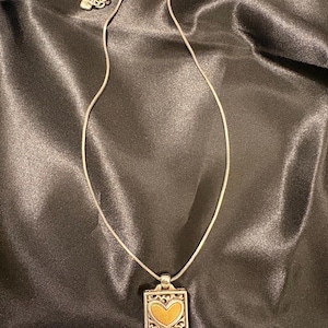 May include: Silver necklace with a square pendant. The pendant has a gold heart design within an ornate silver border. The necklace is displayed on a dark, satin-like fabric.