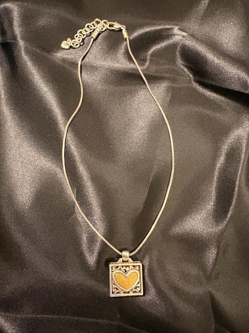May include: Silver necklace with a square pendant. The pendant has a gold heart design within an ornate silver border. The necklace is displayed on a dark, satin-like fabric.