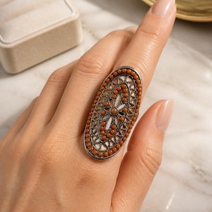 May include: An oval-shaped ring with a silver-colored band. The ring features a detailed design with small orange beads around the perimeter and a central floral pattern. The ring is displayed on a finger.
