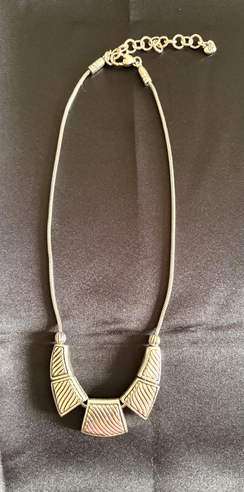 May include: Silver-toned necklace with a geometric pendant. The necklace features a chain and a central pendant composed of three connected shapes. The shapes have a textured, striped design.
