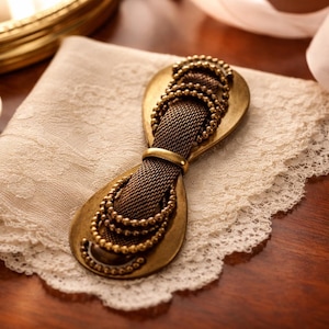 May include: A bronze-coloured metal hair clip with a mesh centre and beaded details. The clip sits on a cream lace doily, with a small dish of pins and a gold-rimmed mirror in the background.