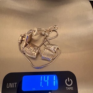 May include: Silver-toned bracelet with two decorative square charms, each with intricate etched designs. The bracelet is on a digital scale, displaying 1.41 oz. The scale is a Pampered Chef brand.