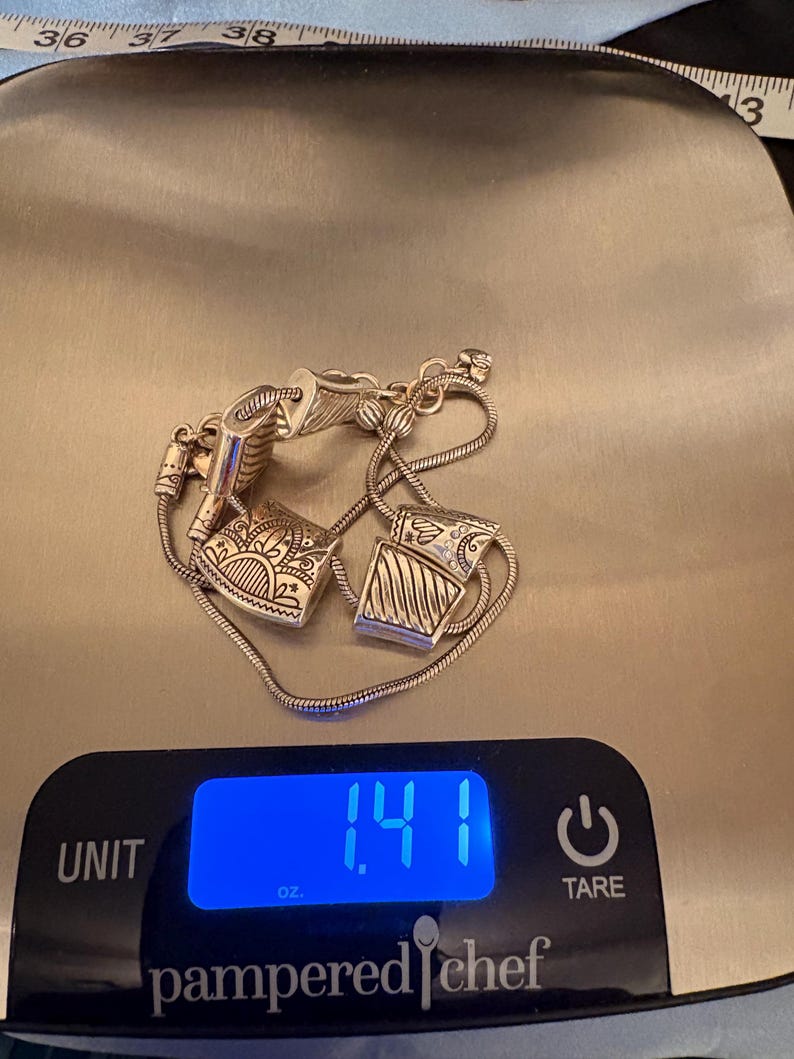 May include: Silver-toned bracelet with two decorative square charms, each with intricate etched designs. The bracelet is on a digital scale, displaying 1.41 oz. The scale is a Pampered Chef brand.