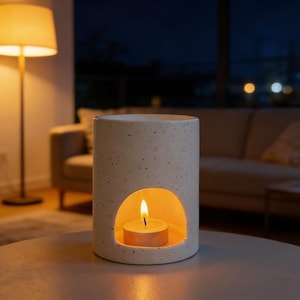 Puede incluir: Un portavelas cilíndrico, moteado, de color blanquecino con una abertura redondeada. Una vela de té encendida brilla en el interior, creando una luz cálida. La pieza decorativa está sobre una mesa.