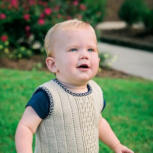 May include: A young child wearing a beige knitted waistcoat with a navy blue trim and matching shorts with an anchor print. The child is standing on a green grassy lawn and looking up.