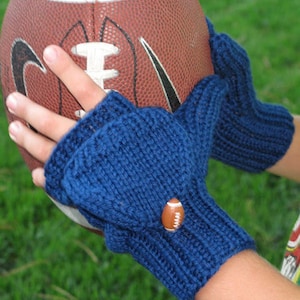 Op de afbeelding: Blauwe gebreide vingerloze handschoenen met een knoop in de vorm van een voetbal. De handschoenen zijn ontworpen om te worden gedragen tijdens het vasthouden van een bruine leren voetbal. De achtergrond is groen.