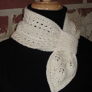 May include: Cream-colored knitted scarf with a leaf-shaped end. The scarf is displayed on a black background. The scarf has a textured pattern and is made of a soft yarn. The scarf is a winter accessory.