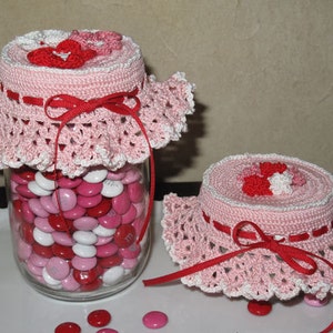 May include: Two crocheted jar covers with pink and white yarn and red ribbon accents. The covers are decorated with small crocheted flowers. The larger cover is on a glass jar filled with pink, white, and red candy.